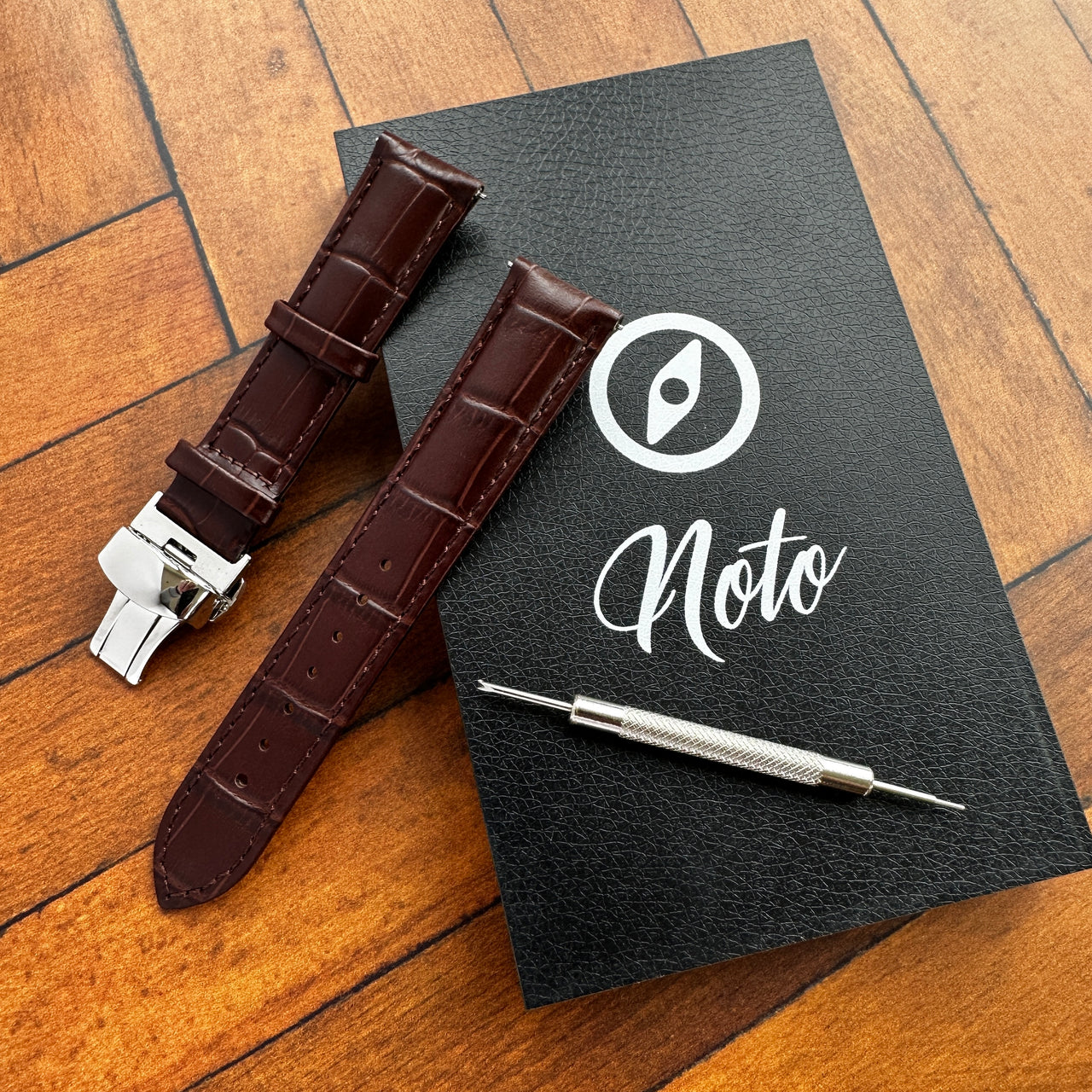 Brown watch strap with silver clasp on a black leather notebook with 'Noto' branding.