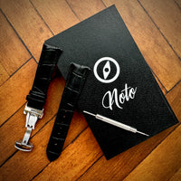 Thumbnail for Black leather watch strap with tool on a black notebook with 'Noto' branding on a wooden surface